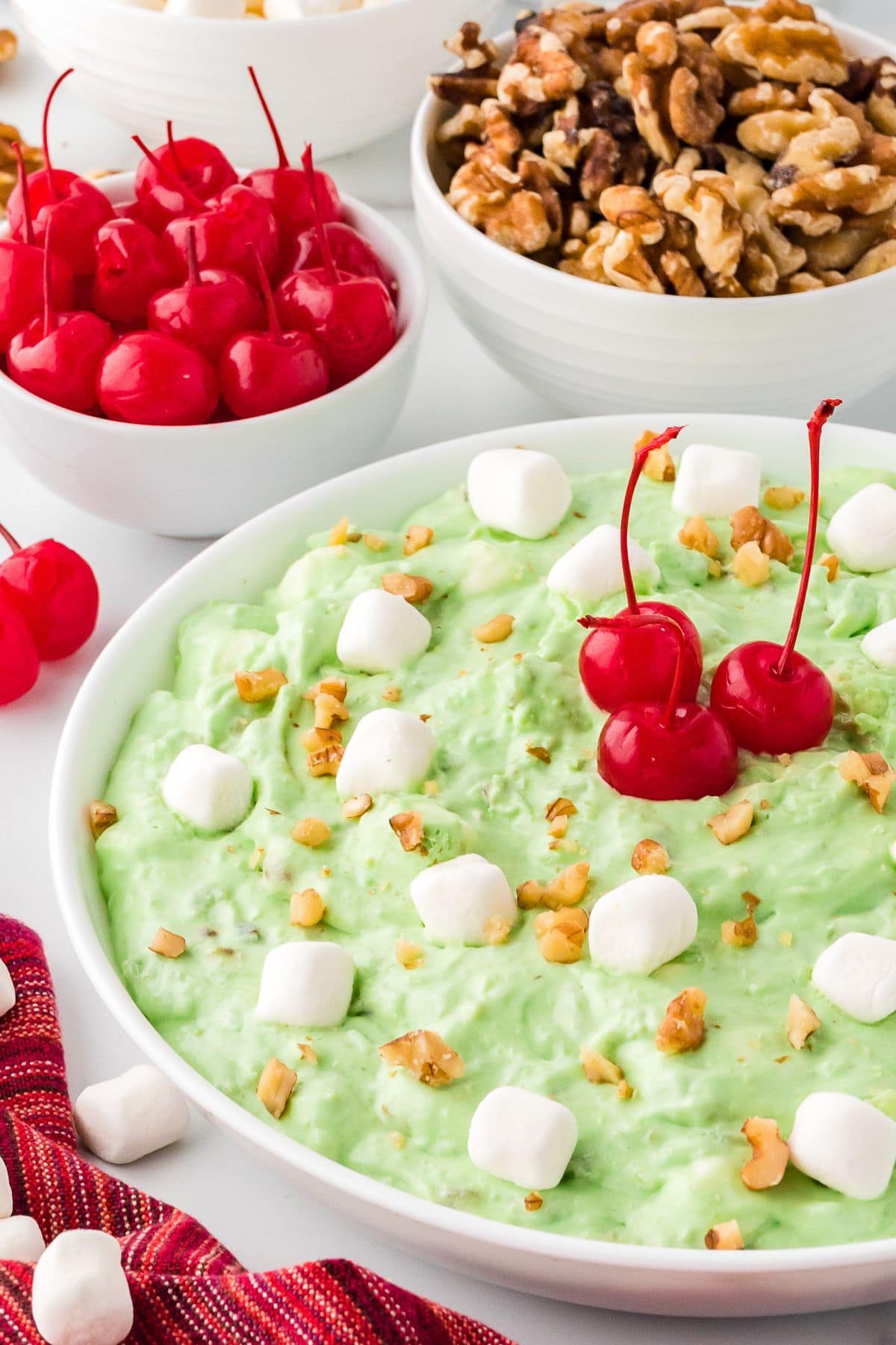 Watergate salad served on a white bowl garnished mini marshmallows, cherries and chopped walnuts.