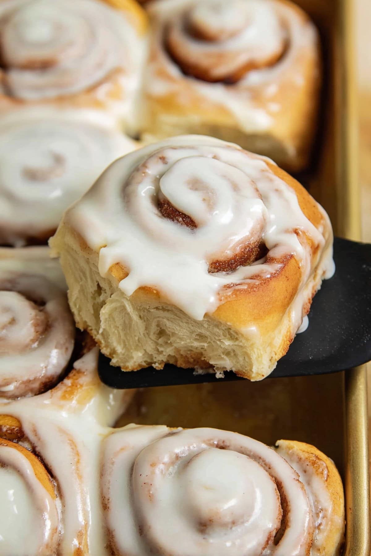 A freshly baked cinnamon roll topped with creamy icing, lifted from a pan with a spatula.