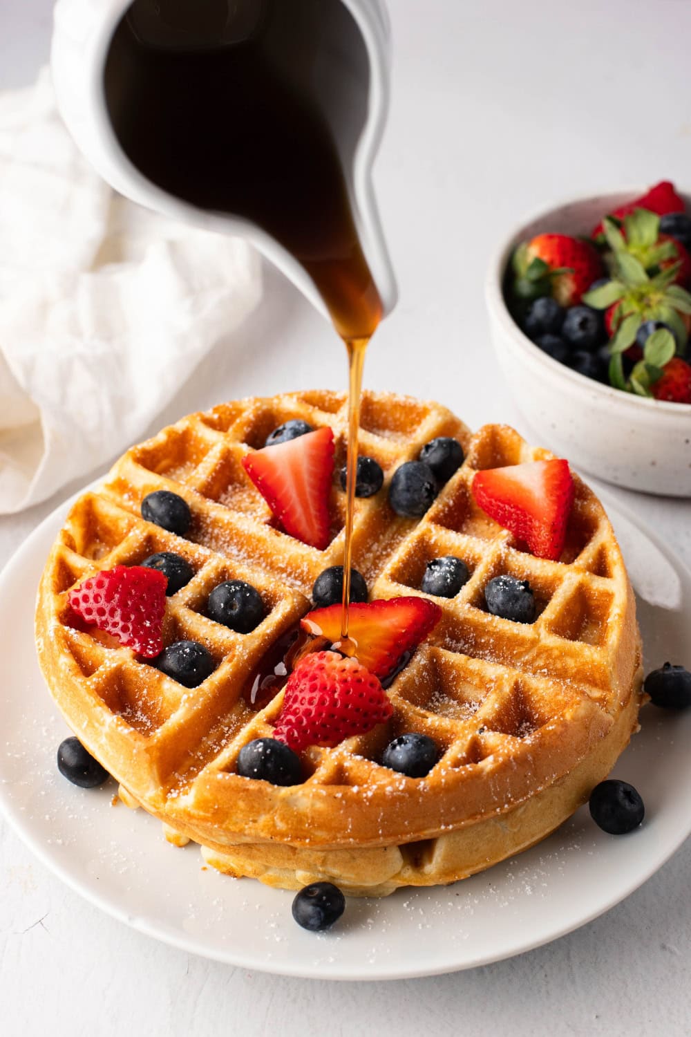 Maple syrup from a ceramic pitcher poured over a stack of waffle in a white plate with berries and strawberry garnish.