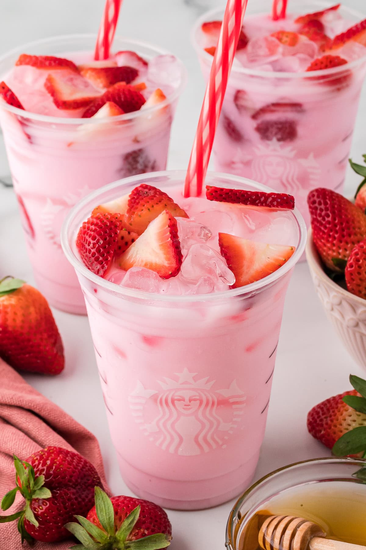 Three Starbucks Pink Drinks in clear Starbucks cups, garnished with fresh strawberry slices.