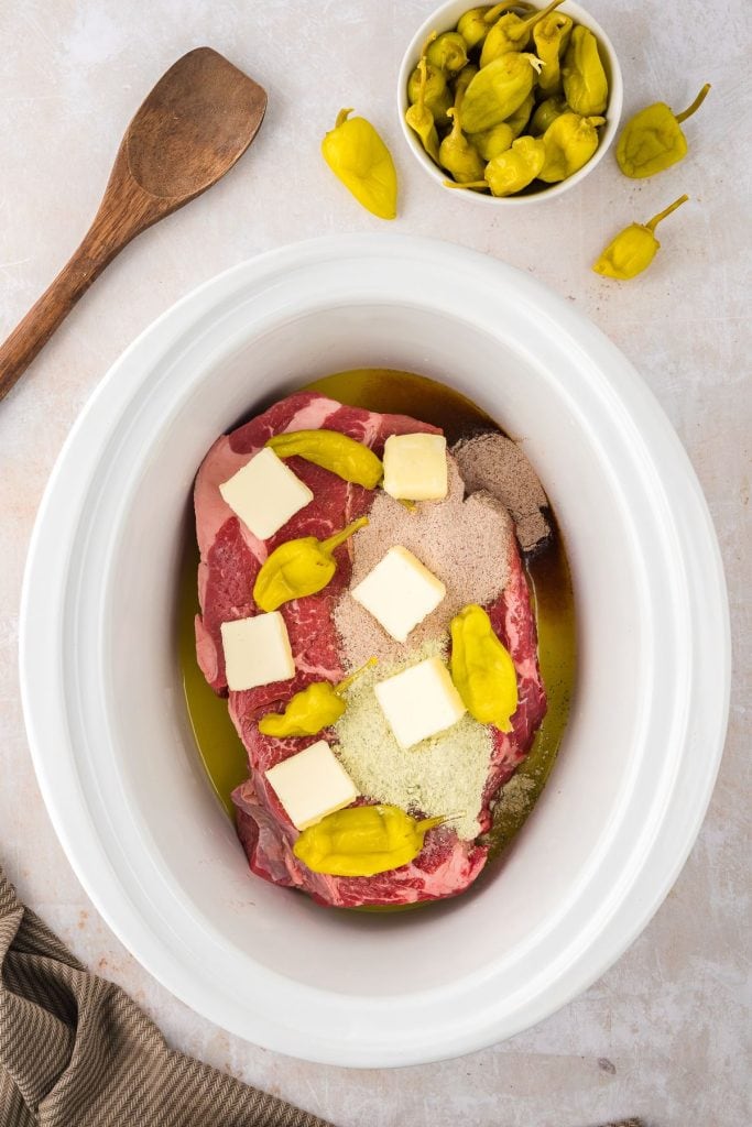 Whole raw chuck roast in a slow cooker with cubed butter, gravy mix, dry ranch seasoning, pepperoncini juice, and pepperoncini peppers, top view