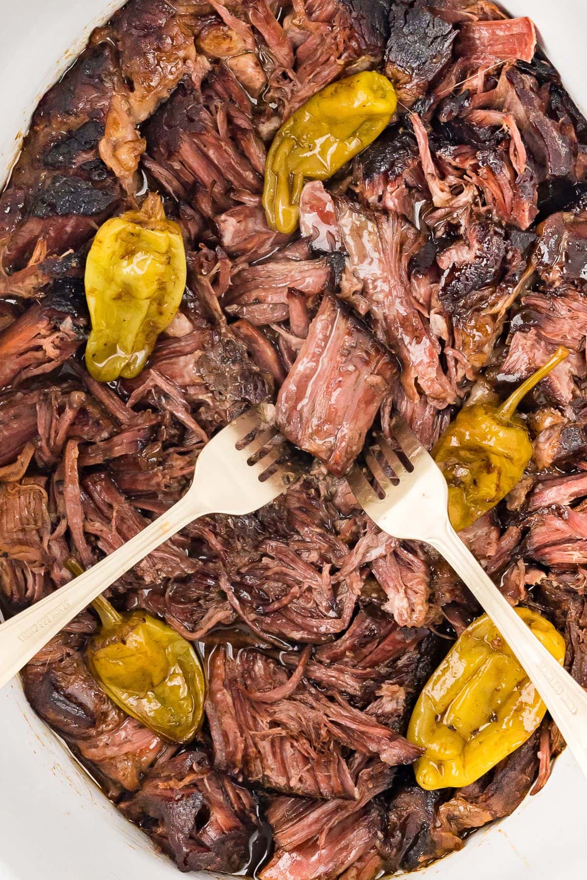 Tender and juicy Mississippi Pot Roast inside a slow cooker with pepperoncini peppers being pulled apart by two forks, close up, top view