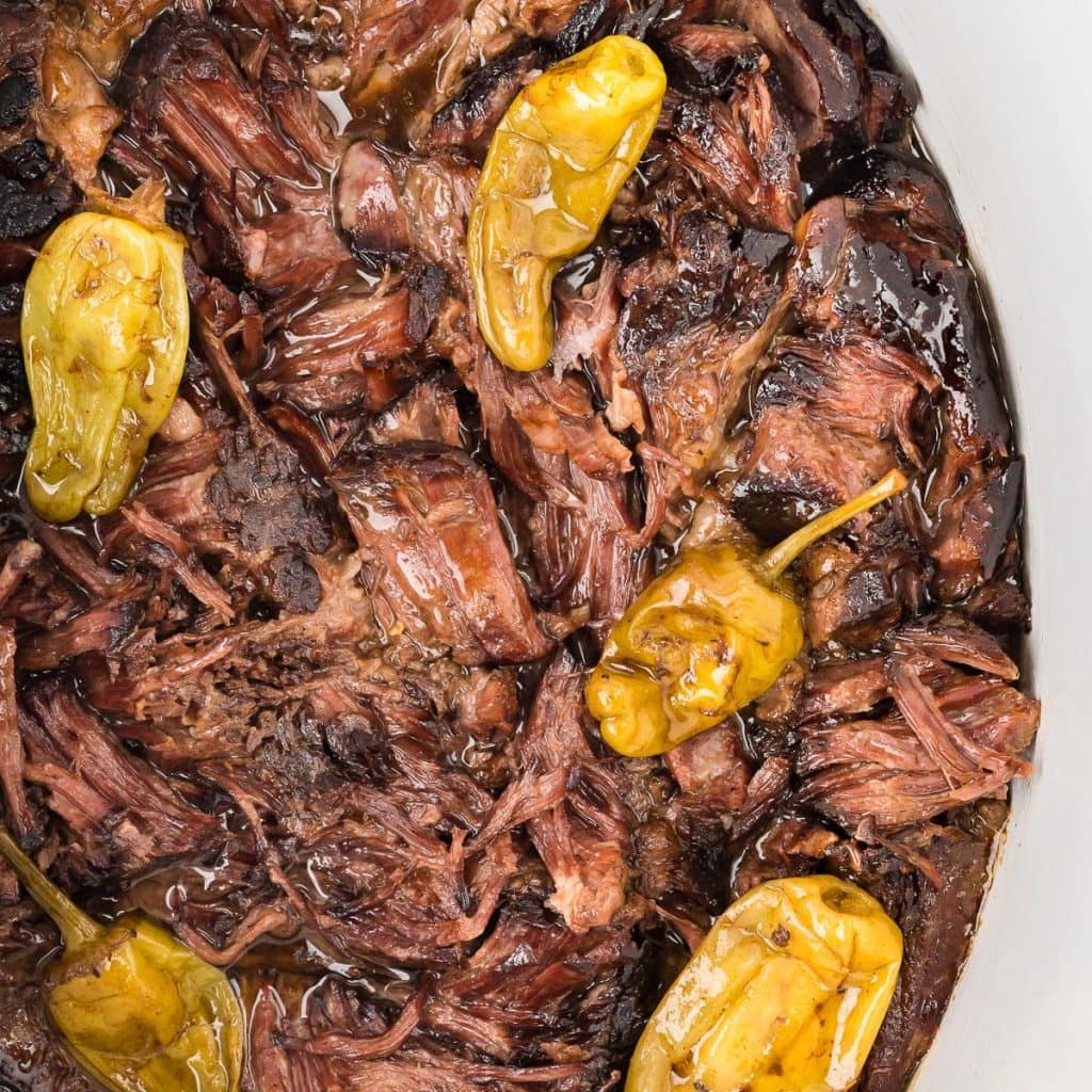 Fork-tender shredded pot roast in gravy with pepperoncini peppers in a slow cooker pot, close up, top view. 
