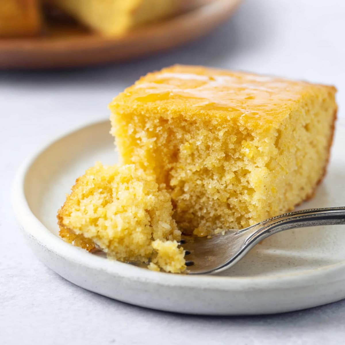 A slice of moist cornbread on a white plate with a fork holding a small bite in front.