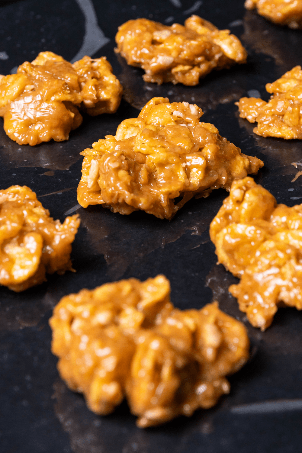 Sticky and glazed peanut butter cornflake cookies arranged on a black surface. 