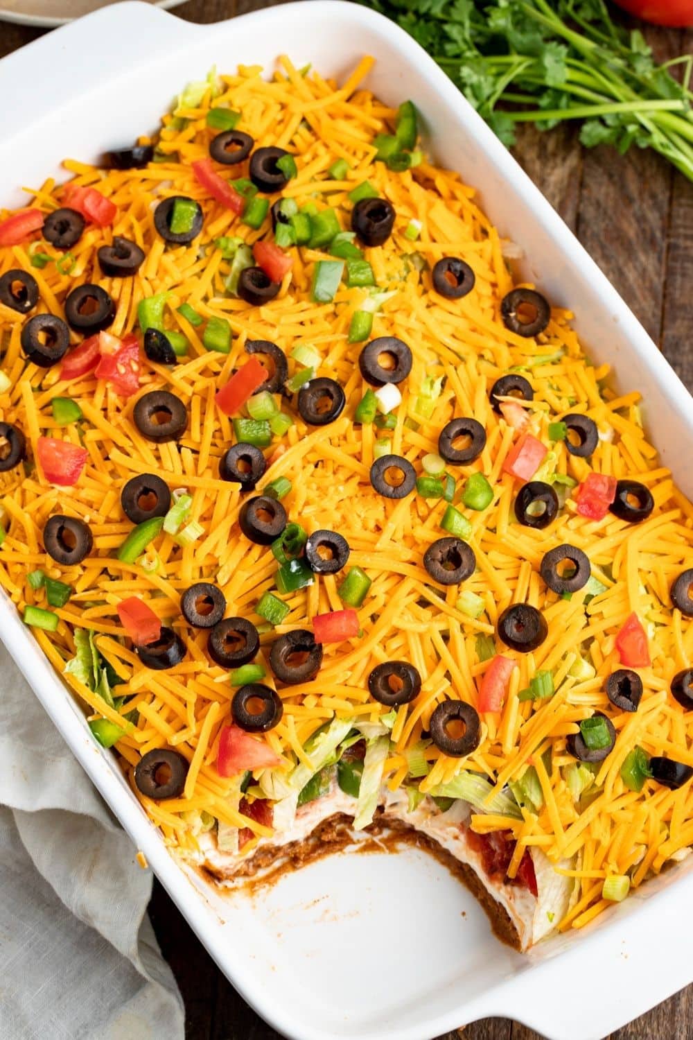 Seven-layer taco dip made with cream sauce, beef, veggies, cheddar cheese, black olives and pepper served on a white casserole. 