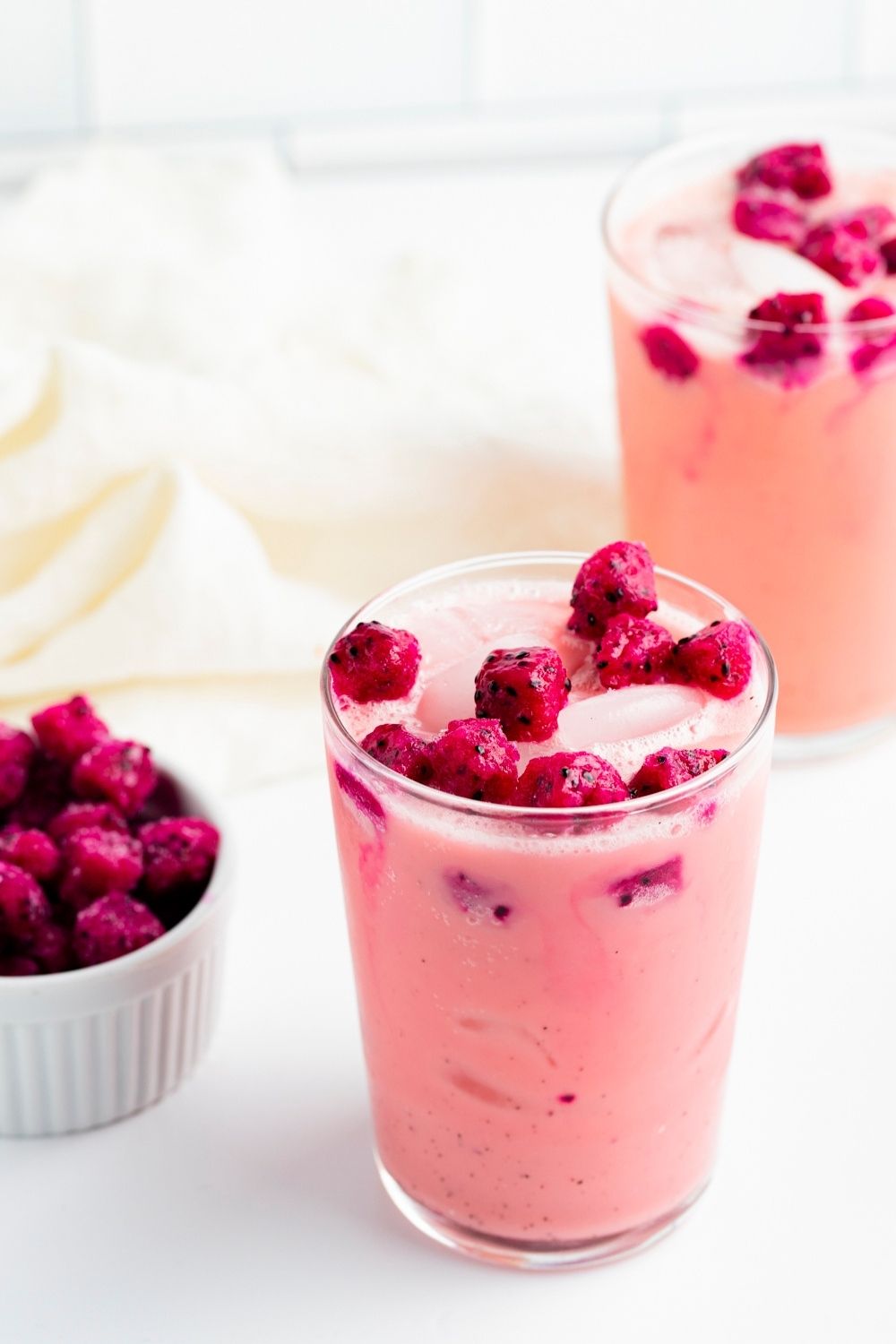 Starbucks Dragon Drink (Copycat Recipe) featuring Two Glasses of Homemade Starbucks Pink Dragon Drink in a Tall Glass with a Dish of Fresh Dragon Fruit Cubes to the Side