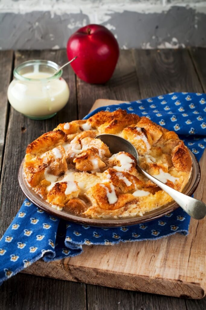 Biscuit bread pudding with vanilla sauce in a bowl on wooden table with apple and blue cloth