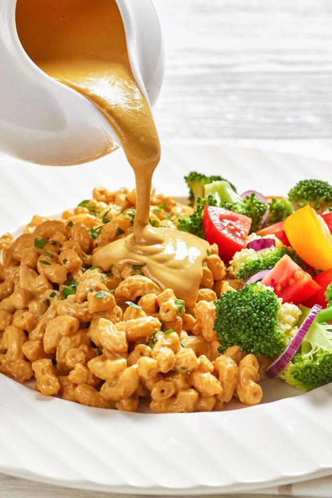 25 Easy Nutritional Yeast Recipes (How To Cook With Nooch). Photo shows jug Pouring Nutritional Yeast Sauce over vegan Mac and Cheese with Vegetables
