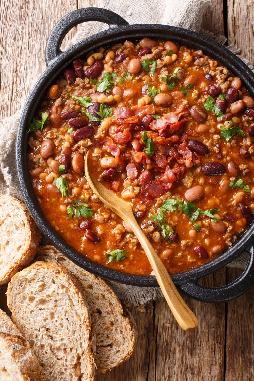 Baked lima and pinto beans with tomato soup base and ground beef on a stone pot garnished with chopped bacon and parsley leaves.