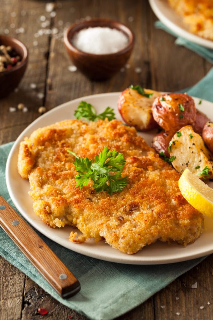Wiener Schnitzel served on a white plate with roasted potatoes and a lemon wedge.