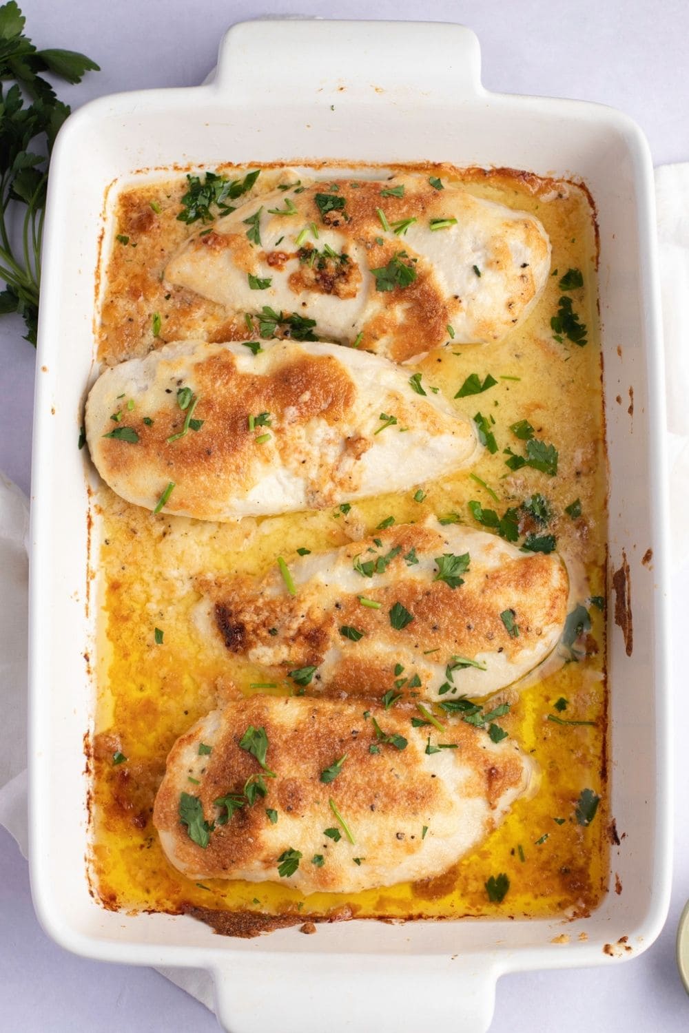 Chicken breast with a cheesy sauce garnished with chopped parsley leaves on a casserole dish.