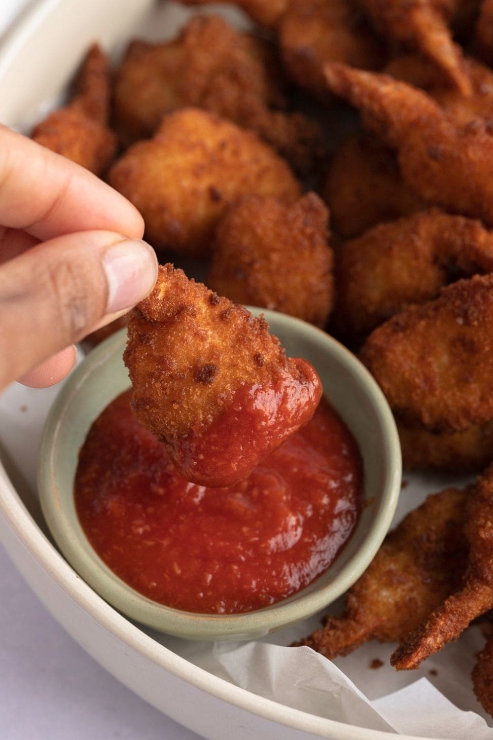 Hand Dipping Breaded Air Fryer Shrimp in a Bowl of Red Cocktail Sauce with More Shrimp on a Dish 