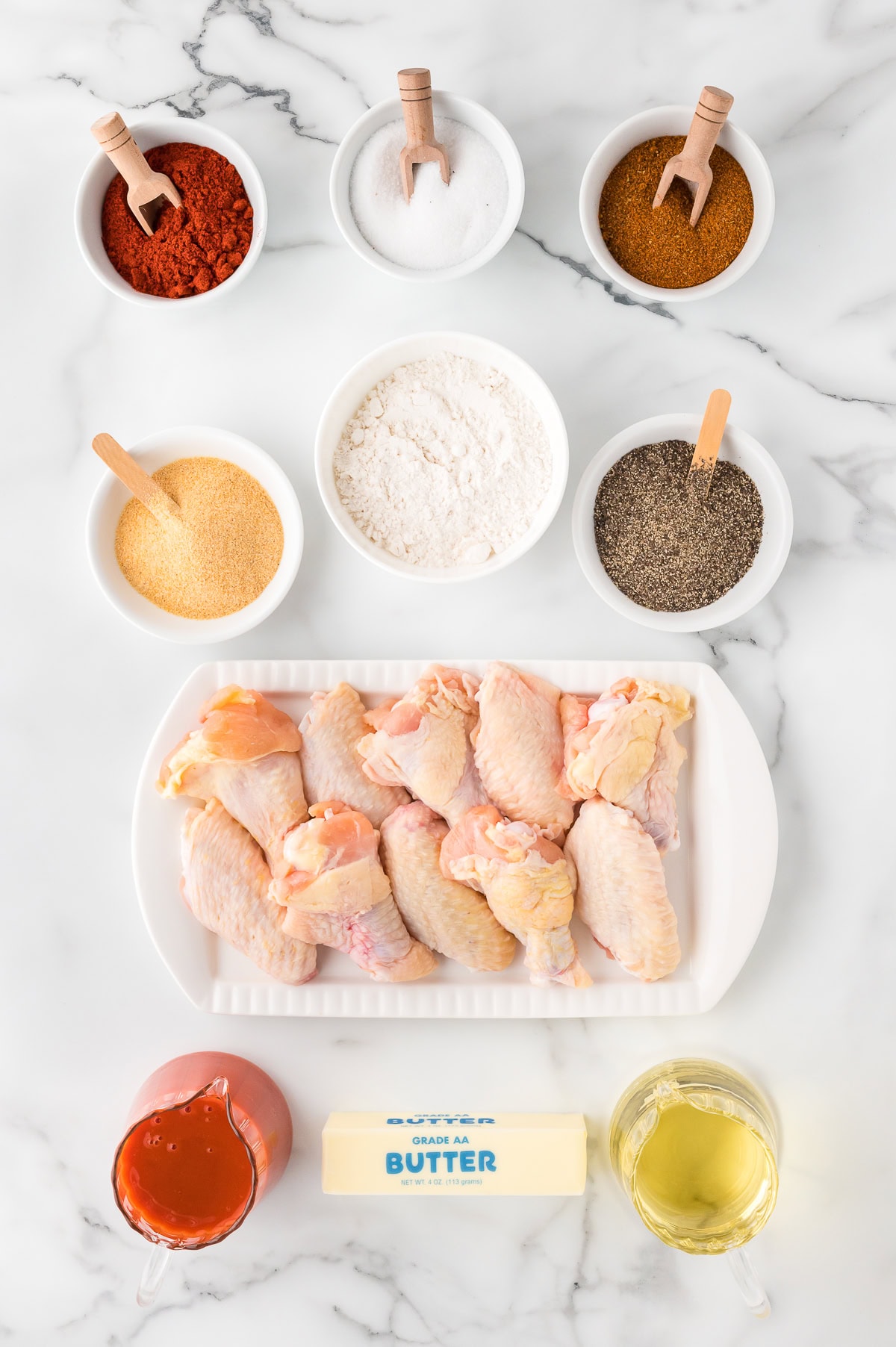 Buffalo Chicken Wings ingredients: Raw chicken wings, flour, paprika, garlic powder, salt, black pepper, cayenne pepper, oil, hot sauce, a stick of butter, and brown sugar, top view