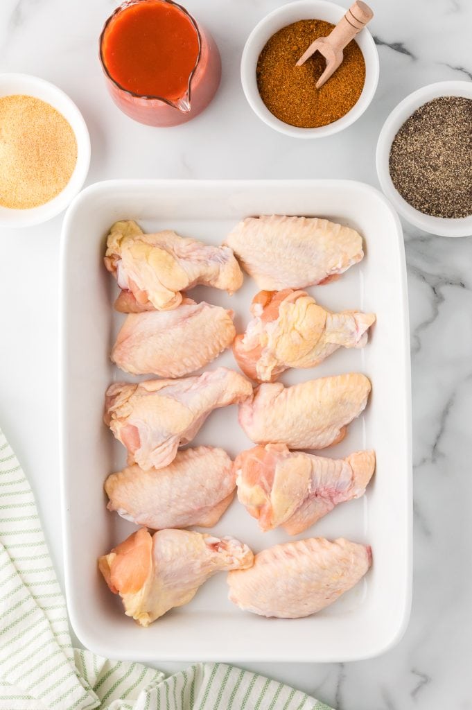 Raw chicken wings in a baking dish with spices and buffalo sauce on the side, top view