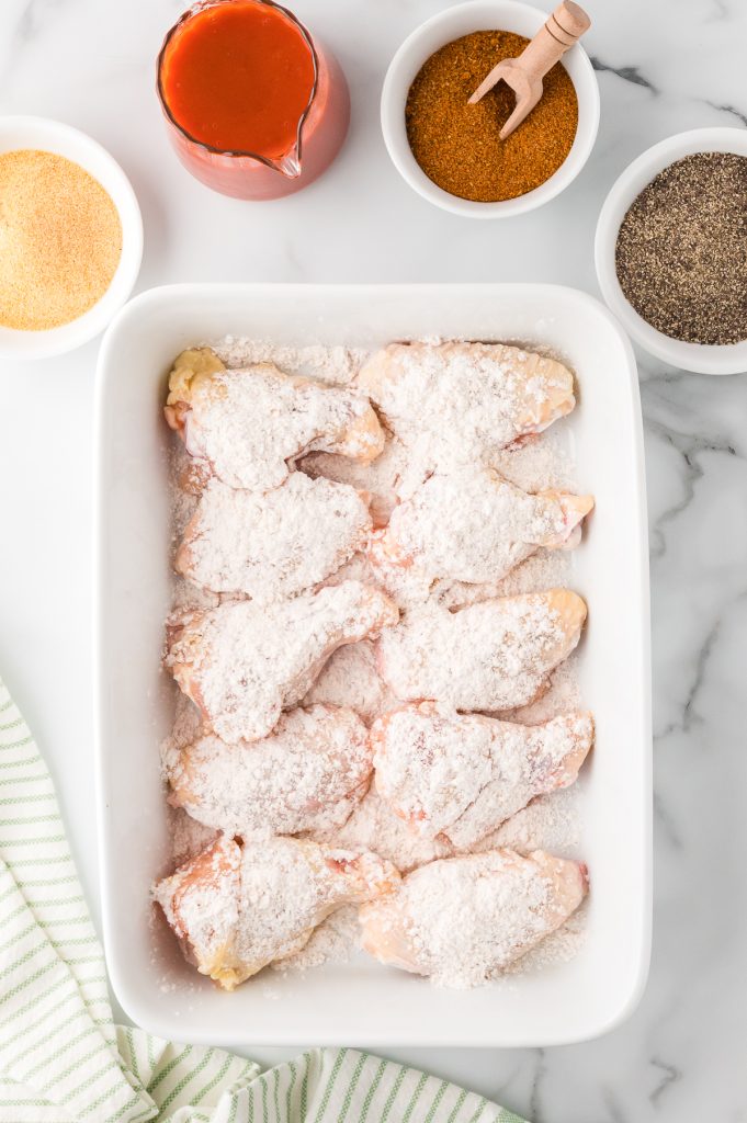 Raw chicken wings coated with spiced flour mixture in a baking dish with more spices and buffalo sauce on the side, top view.