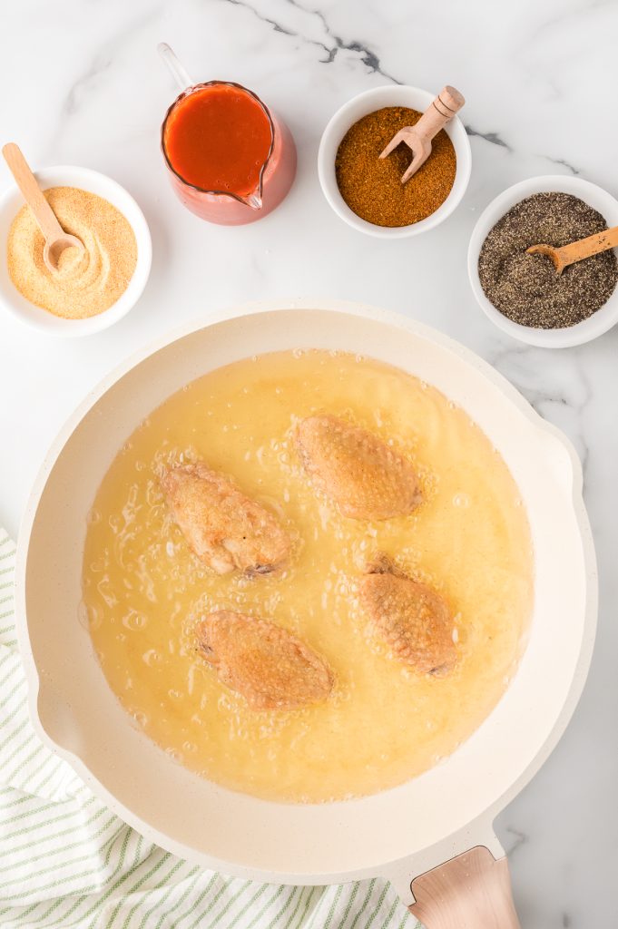 Fried chicken wings in a deep skillet with buffalo sauce and spices on the side, top view.