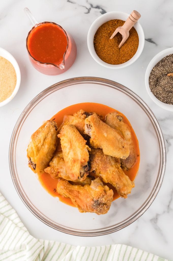 Crispy chicken wings in a glass bowl with buffalo wing sauce and extra sauce and spices on the side, top view.