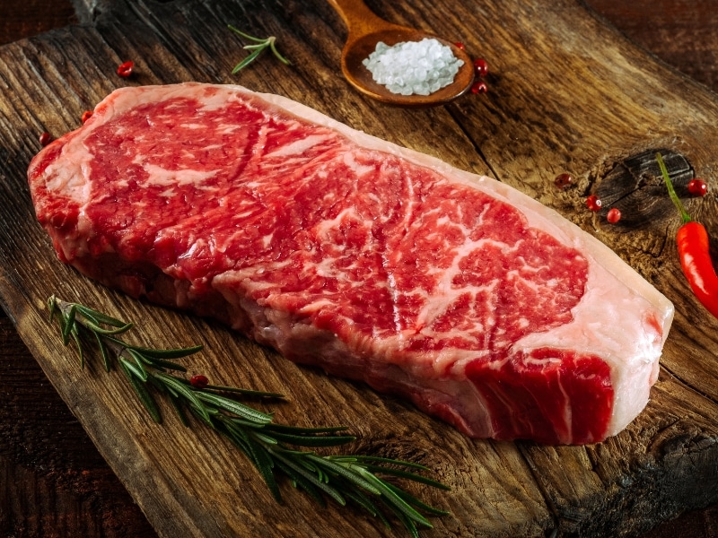 Raw Strip Steak on a Wooden Cutting Board with Chili and Rosemary
