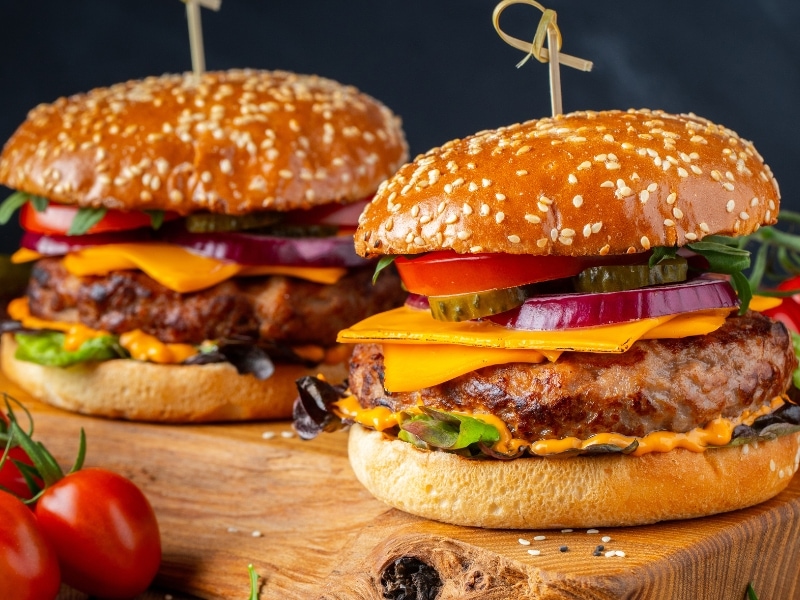 Two Burgers on a Wooden Cutting Board with Onion Tomato Cucumber and Cheese