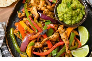 Top View of Chicken Fajitas with Lime Wedges and Guacamole on a Plate on a Wooden Cutting Board with Tortillas