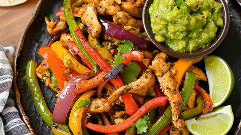 Top View of Chicken Fajitas with Lime Wedges and Guacamole on a Plate on a Wooden Cutting Board with Tortillas