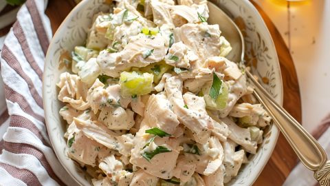 Top View of Ina Garten's Chicken Salad in a White, Patterned Bowl with a Spoon on a Wooden Cutting Board on a White Table with a Kitchen Towel