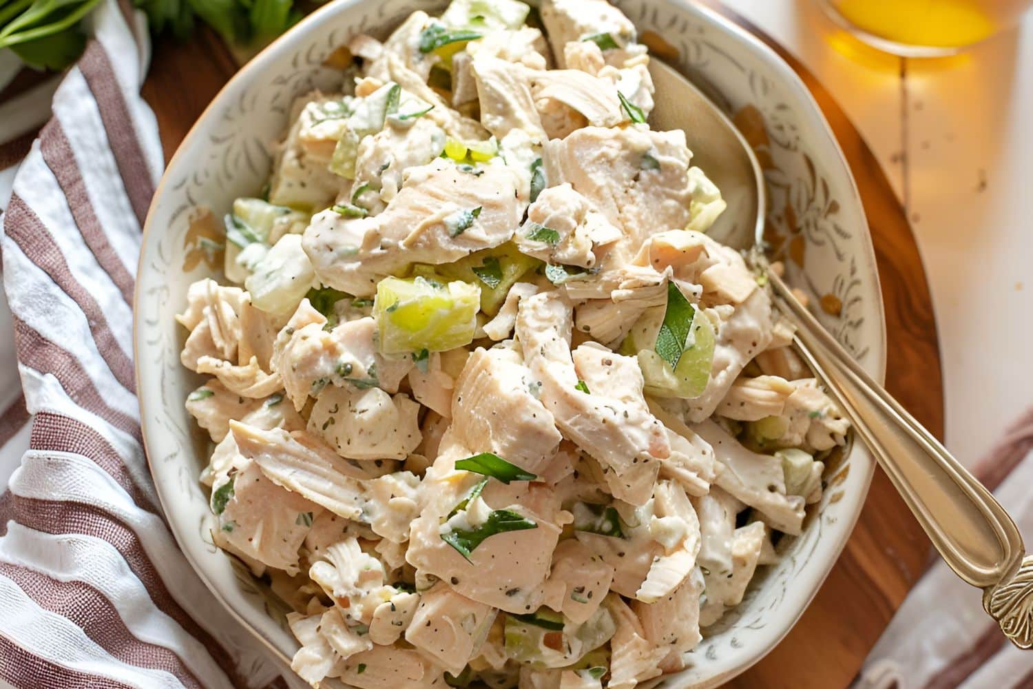 Top View of Ina Garten's Chicken Salad in a White, Patterned Bowl with a Spoon on a Wooden Cutting Board on a White Table with a Kitchen Towel