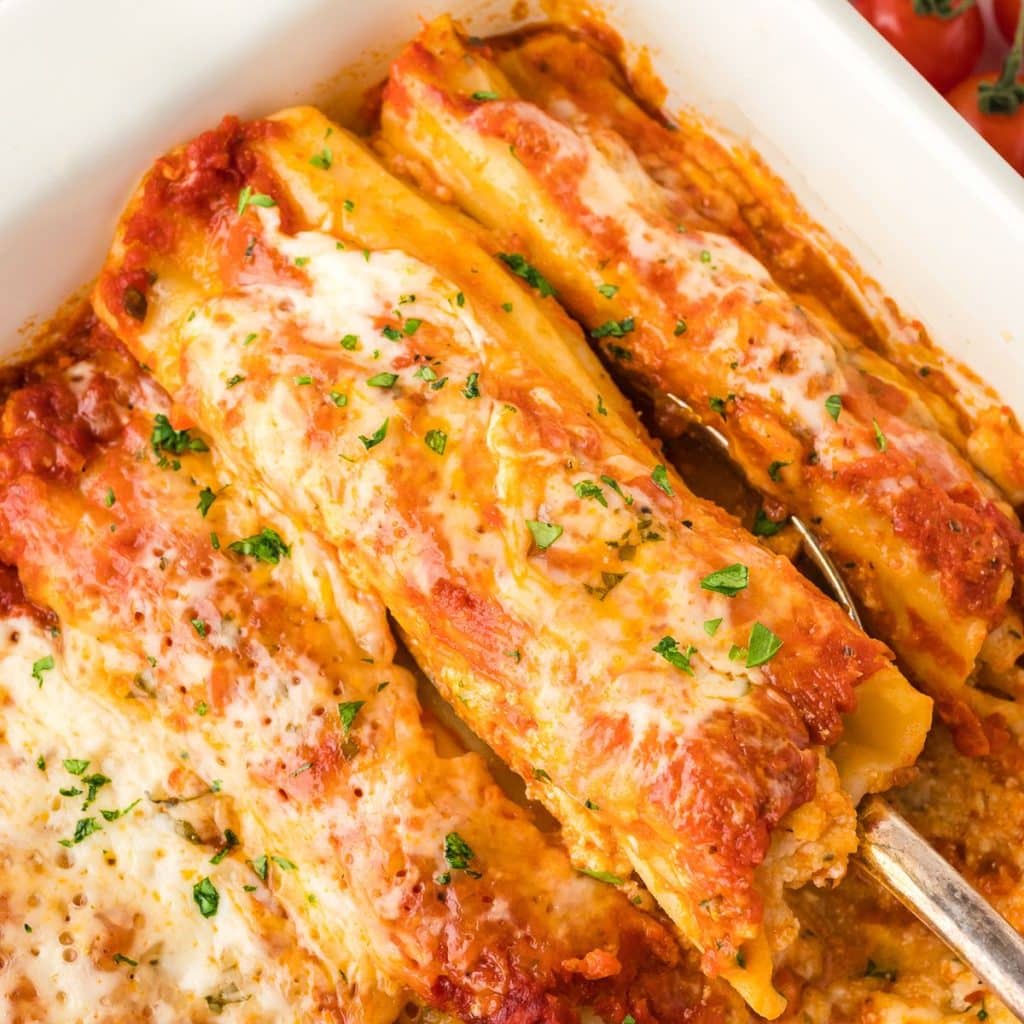 A metal spoon lifting a serving of no-boil cheese manicotti from a white baking dish, close up