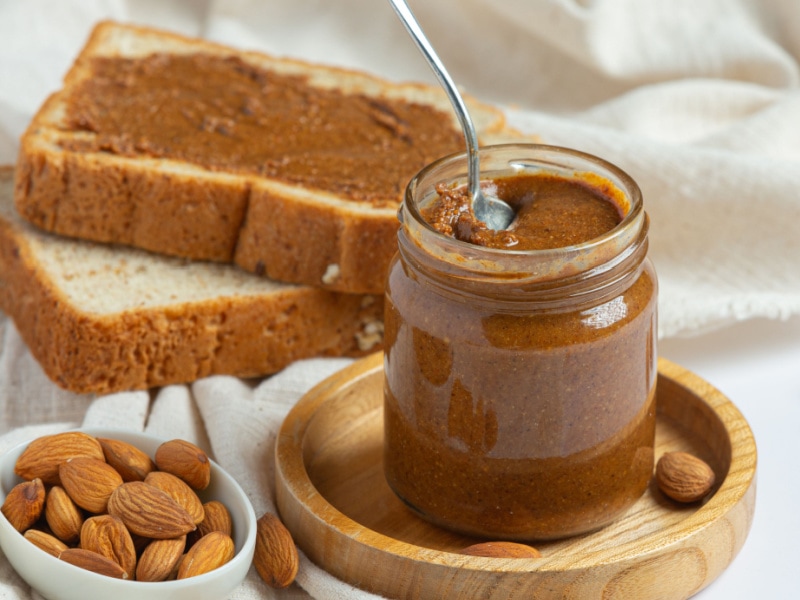 Almond butter in a glass jar with a bowl of almonds and sliced bread