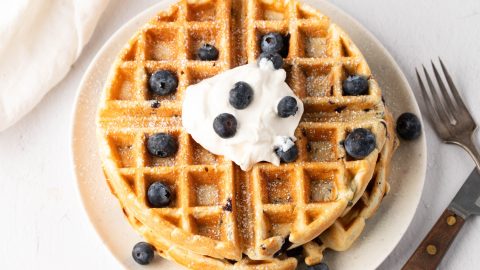 Stack of blueberry waffles with whipped cream on top served in a white plate.