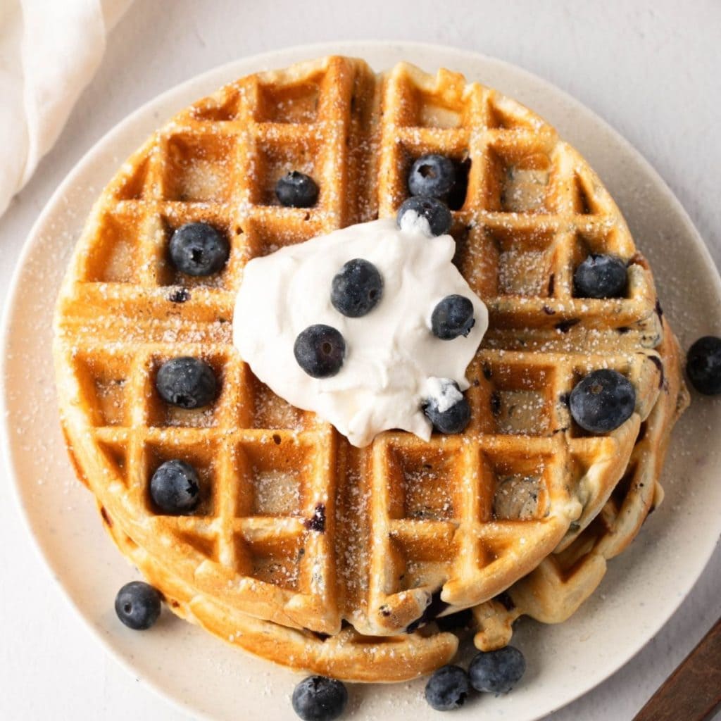 Waffles with blueberries and whipped cream on top dusted with powdered sugar served on a white plate.