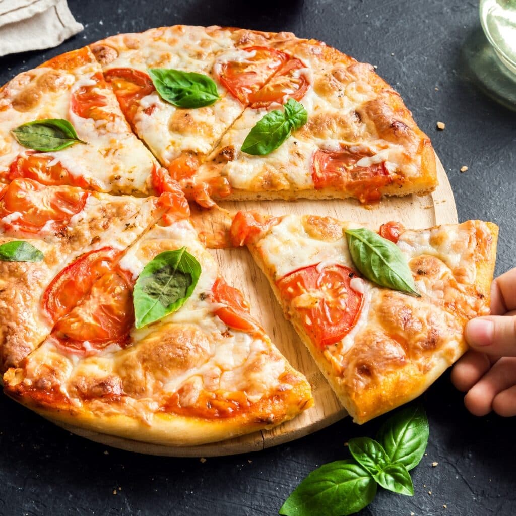 A Hand Holding A Slice Of Cheesy Pizza Margherita With Tomatoes, Garnished With Fresh Basil Leaves. 