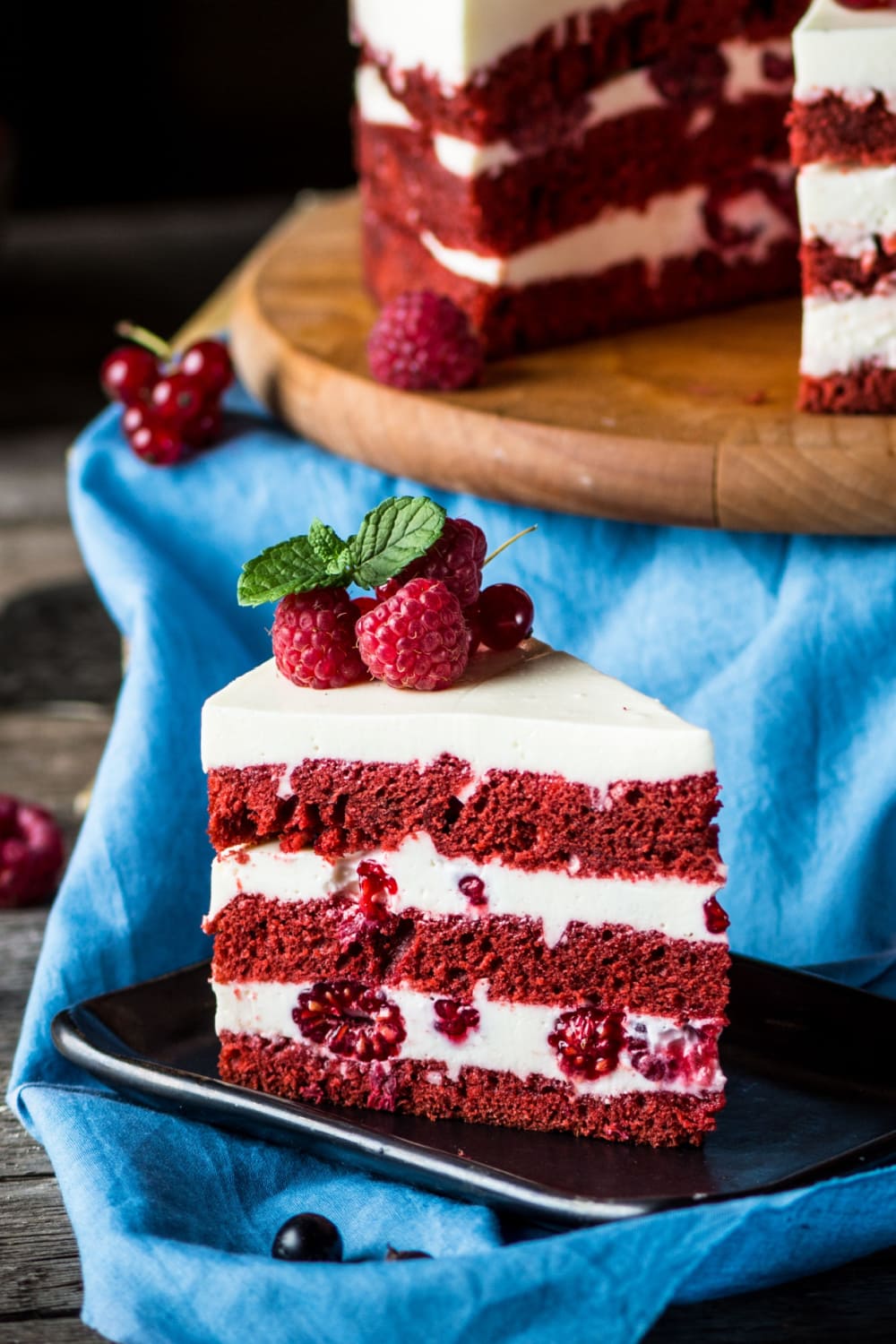 Slice of Red Velvet Cake Layered With Cream and Moist Cake, Topped With Fresh Raspberries Served on a Black Square Plate
