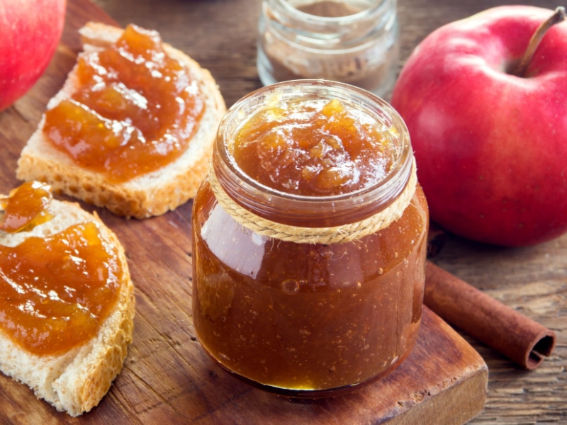 Apple butter in a small jar with two slices of bread with filling, cinnamon sticks and fresh whole apple on side. 