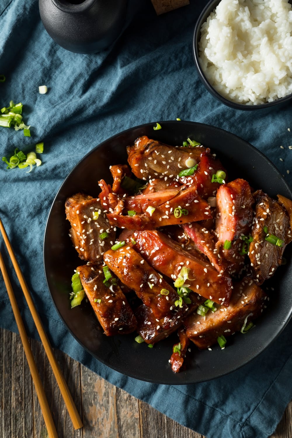 Bowl of White Rice and Chinese BBQ Pork Ribs Served on a Black Plate Garnished With Sesame Seeds and Chopped Onion Leeks