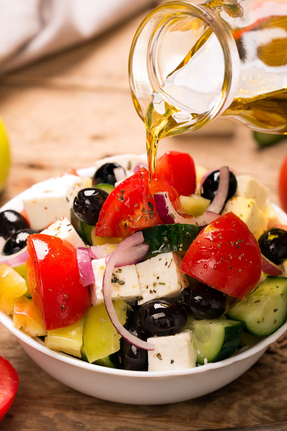 Olive oil pouring over a bowl of Greek salad with olives, feta cheese and tomato slices. 