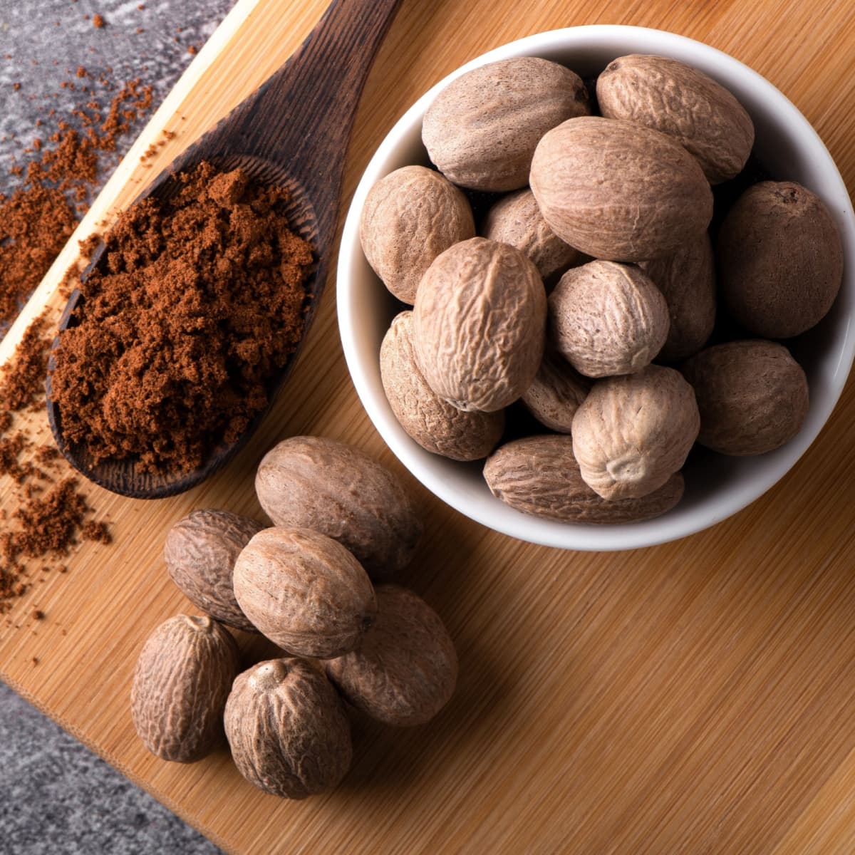 Whole Nutmeg in a White Bowl and Powdered Nutmeg in a Wooden Spoon on A Wooden Plate
