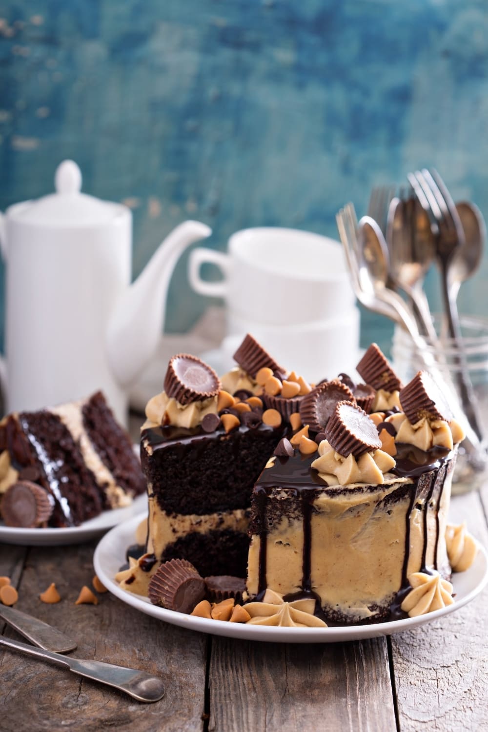 A sliced whole Chocolate Peanut Butter Cake, revealing its layers of chocolate and cream, topped with Reese's and chocolate chips, served on a white plate. 