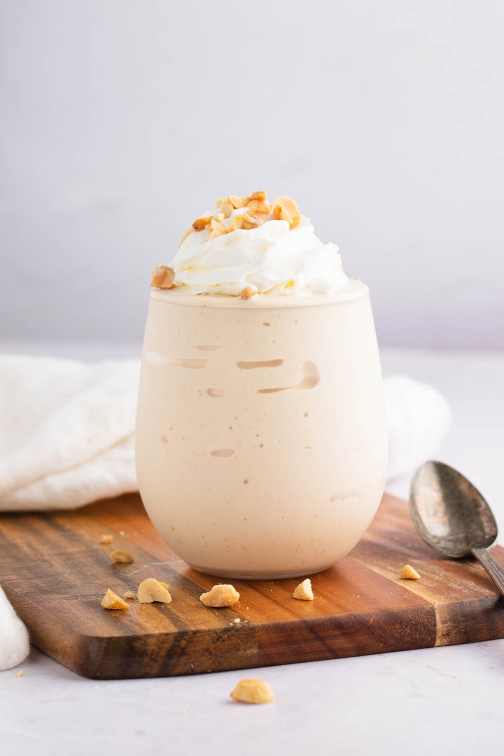Peanut Butter Milkshake Garnished with Nuts  and Whipped Cream on Top of a Wooden Board With a Spoon on Side