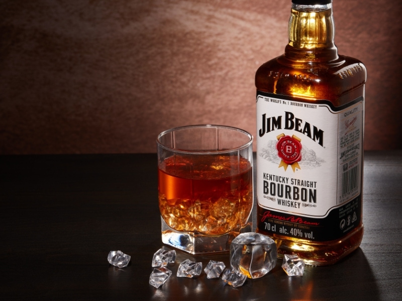 Sebotol wiski bourbon Jim Beam dan Gelas Dengan Minuman dan Es di Meja Kayu 
