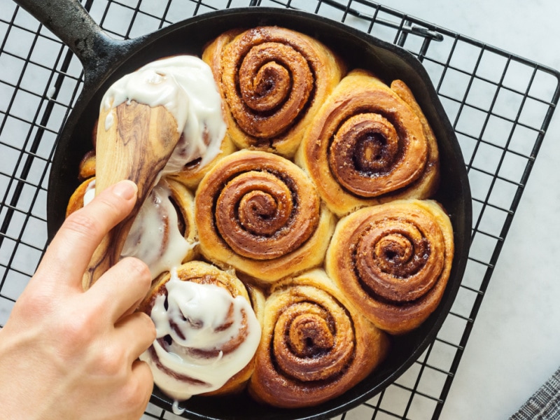 Cinnamon rolls in a skillet with a person spreading glaze on top