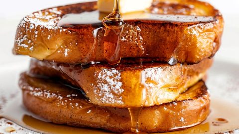 Three Slices of Eggnog French Toast Stacked with Butter, Powdered Sugar, Cinnamon, and Maple Syrup on a White Plate