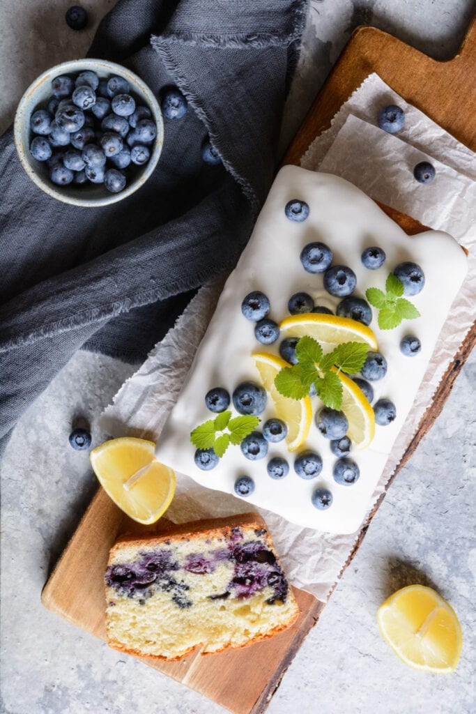 Lemon blueberry bread on a chopping board with fresh blueberrues and lemon wedges, top view