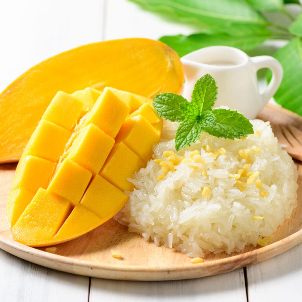 Ripe Mango Served With Scoop of Sticky Rice Garnished with Fresh Basil Leaves on a Wooden Plate