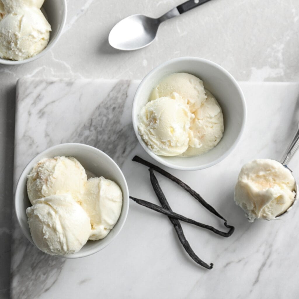 Creamy Vanilla Ice Cream Scoops in Bowl, Sticks of Dried Orchard Seed Pods on a Marble Table