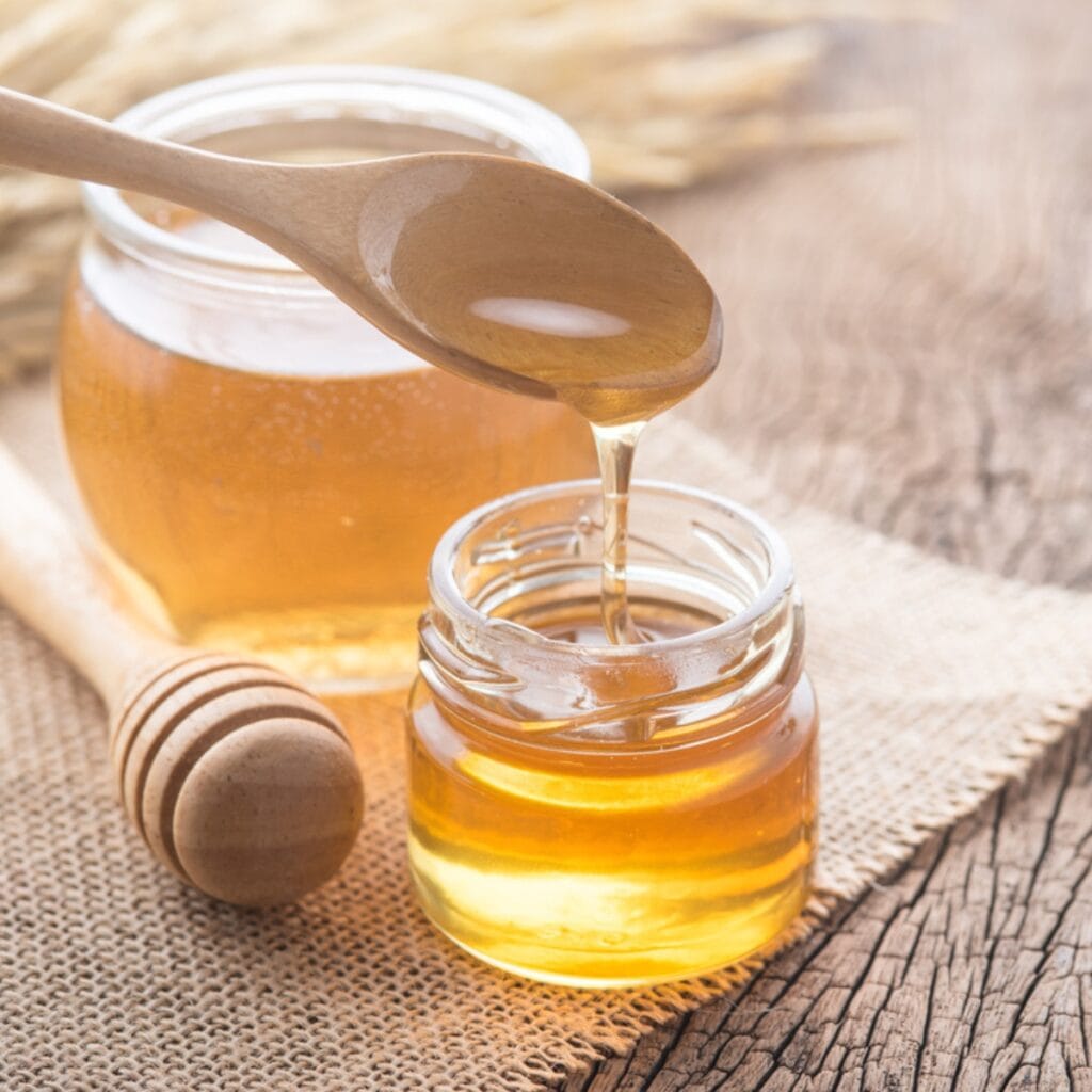Honey Dripping From Wooden Spoon Into Glass Jar, Wooden Dipper on Wooden Table, Larger Jar of Honey in Background

