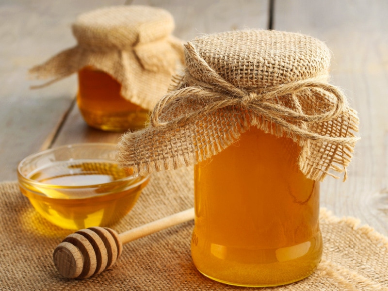 Two Jars of Honey with Cute Burlap Covers, Tied with Jute on a Wooden Table with a Honey Dipper and a Small Glass Bowl of Honey