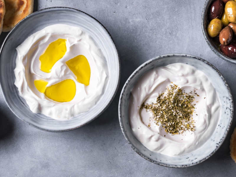 Two Bowls of Labneh Cheese, One Drizzled with Oil and One Topped with Za'atar and a Bowl of Olives in the Background