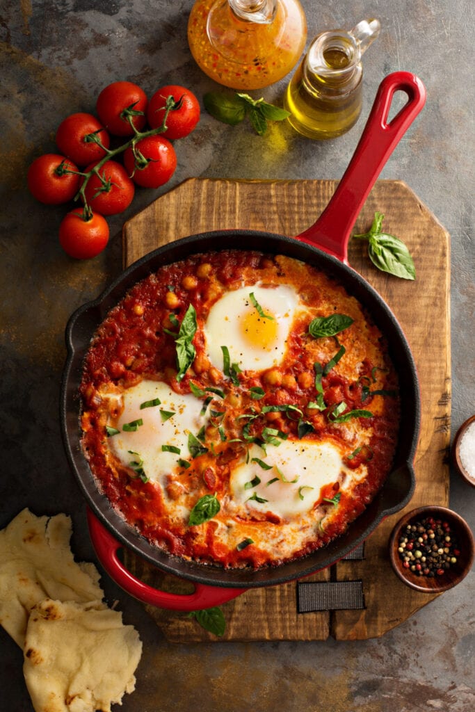 Shakshuka in a Pan on a Wooden Cutting Board with Fresh Tomatoes, Basil, and Peppercorns Around the Board